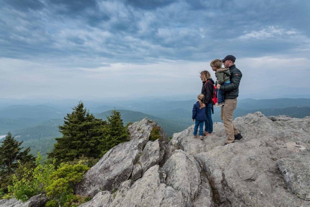 A family of four stands on a rocky mountain edge, overlooking a vast, green landscape under a cloudy sky. An adult holds a child in a carrier, while another adult and child stand nearby, all bundled in jackets.