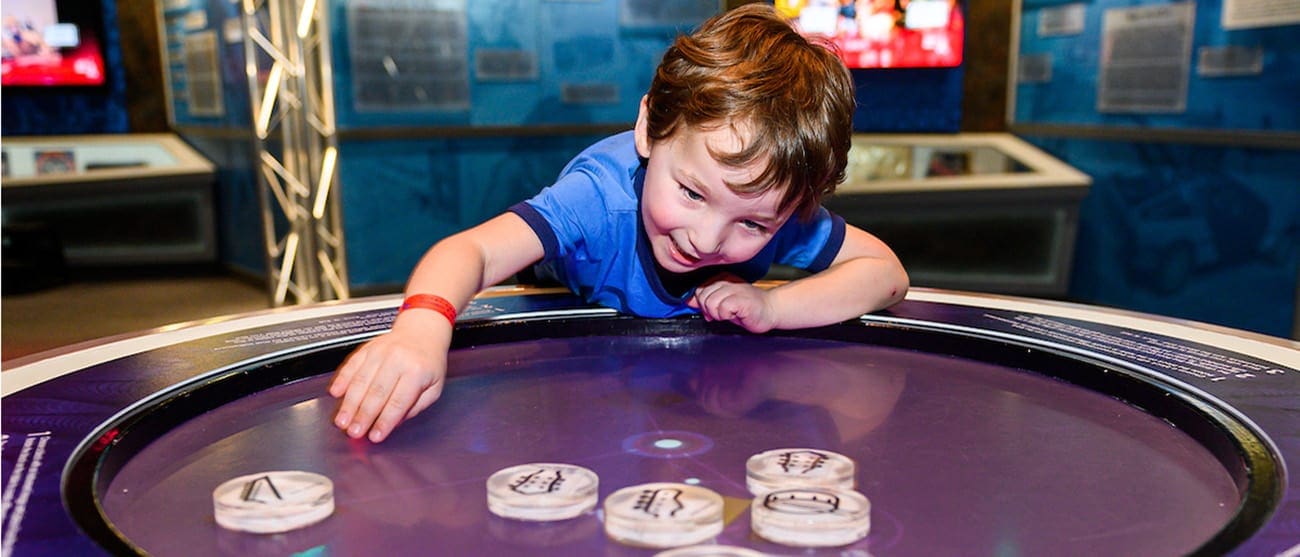 A young boy in a blue shirt excitedly interacts with a circular display table, moving wooden discs featuring various symbols. The background features informative panels and a screen.
