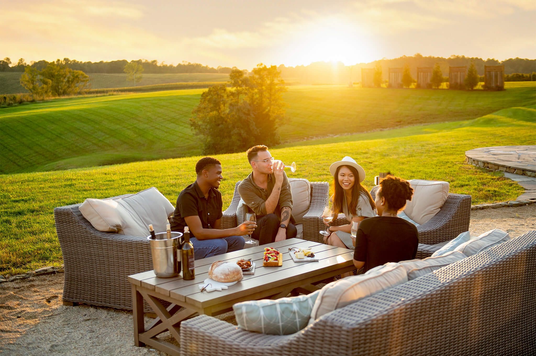 Four people sit on outdoor furniture on a sunny day, enjoying drinks and snacks. They are surrounded by a lush green landscape with a setting sun in the background, creating a warm and inviting atmosphere.