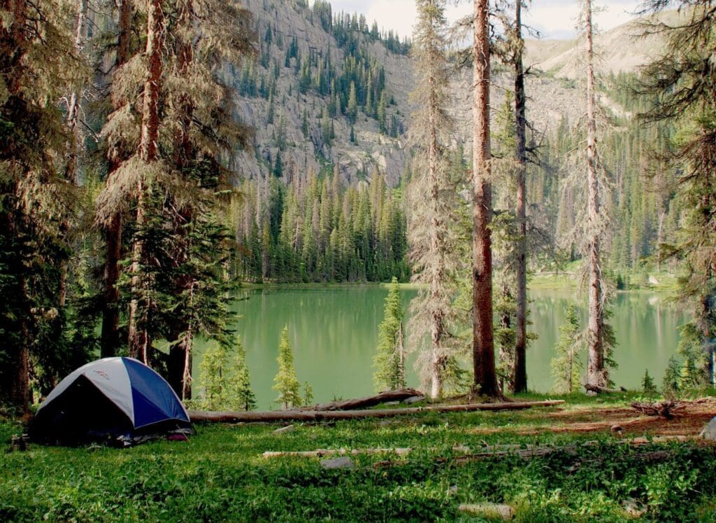 A small tent in a forested area overlooks a serene, green lake surrounded by tall pine trees and distant rocky cliffs. The scene is calm and lush, with scattered logs and soft sunlight filtering through the trees.