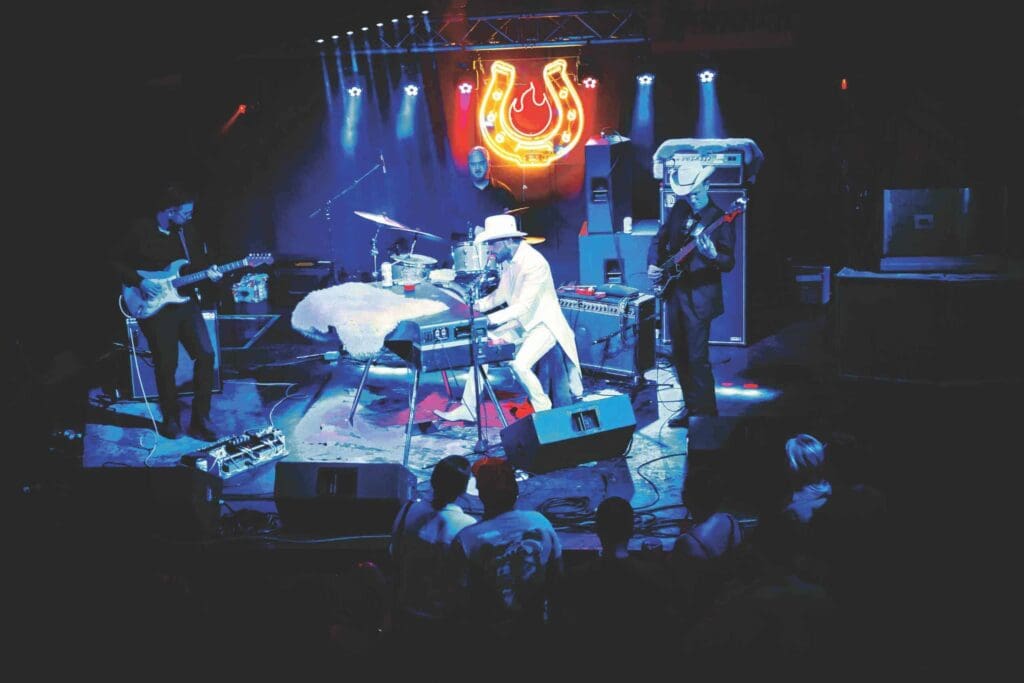A band performs energetically on a dimly lit stage, featuring a keyboardist in a white suit and hat. The backdrop has a red neon horseshoe sign. Onlookers stand in the foreground, enjoying the music.