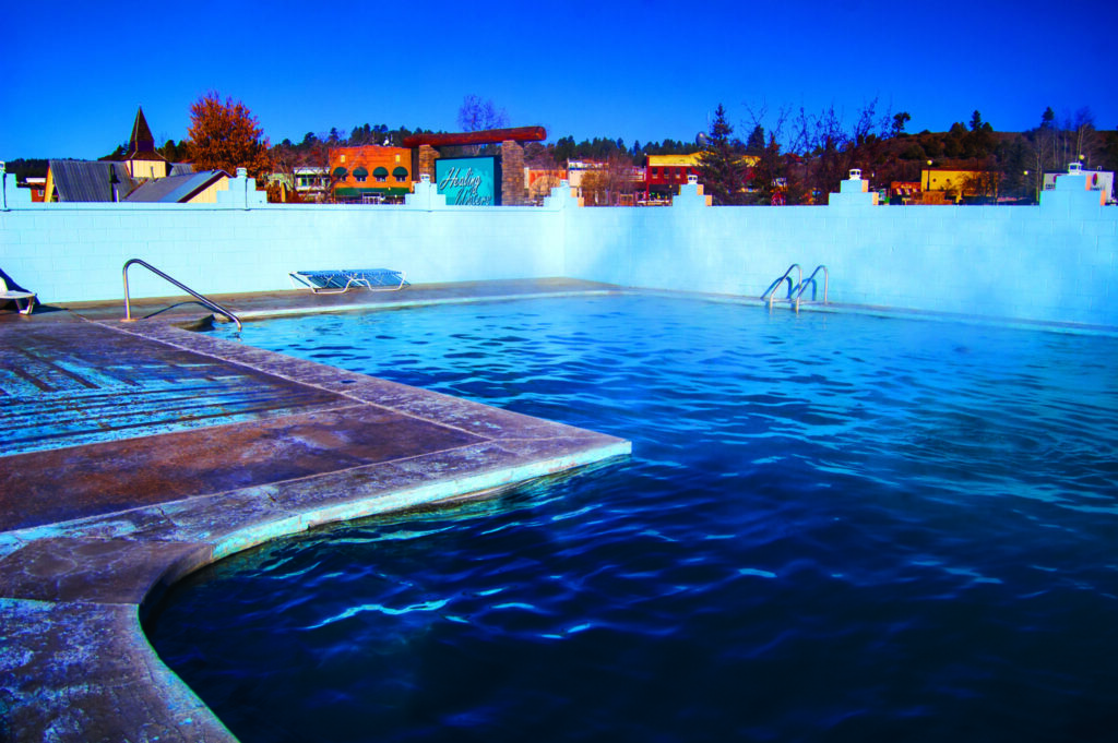 Outdoor swimming pool with steamy water surrounded by a concrete deck. There are two metal pool ladders and a lounge chair nearby. In the background, there are buildings and trees under a clear blue sky.
