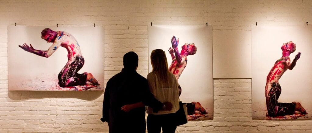 A couple stands closely, admiring three large photographs on a gallery wall. The images depict a person kneeling and covered in vibrant paint, set against a white brick background.