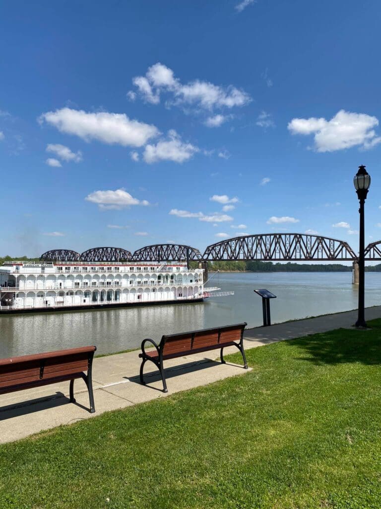 A riverside scene with two empty benches on a grassy area. A paddleboat is docked on the river, and a large iron bridge spans the water in the background under a clear blue sky with scattered clouds. A lamppost stands nearby.