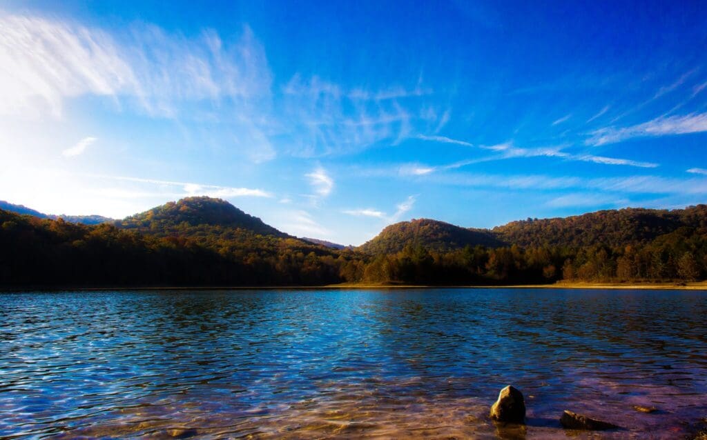 A serene lake reflecting the blue sky is surrounded by tree-covered hills. Wispy clouds stretch across the sky. The foreground features clear, rippling water and a few rocks at the shoreline.