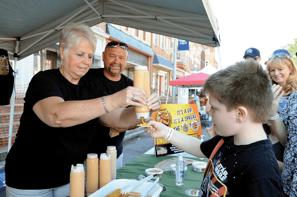 A woman hands a bottle of sauce to a boy at an outdoor food stall. A man stands beside her, smiling. People wait in line behind the boy. The stall has condiments on a table and a sign promoting dips.