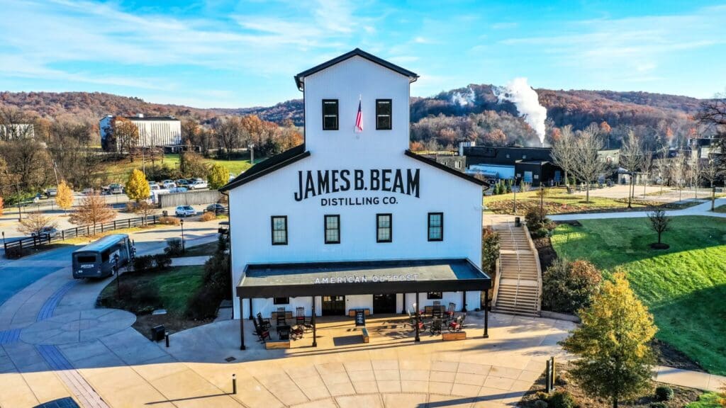 Aerial view of James B. Beam Distilling Co., a white, barn-like building with black accents. Surrounding it are trees and green lawns, a tour bus, and distant industrial structures with smoke. Clear blue sky overhead.
