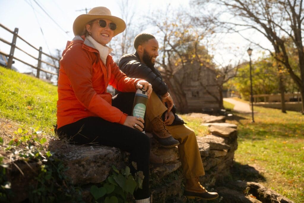 Two people sit on a stone wall outdoors. The person on the left wears a sun hat, sunglasses, and an orange jacket, holding a bottle. The person on the right has a beard and wears a dark jacket. Trees and a path are in the background.