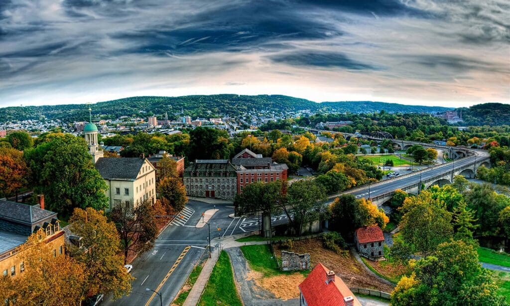 Aerial view of a small town with a mix of historic and modern buildings surrounded by lush greenery and autumn foliage. A bridge stretches across the landscape, with rolling hills and a dramatic, cloudy sky in the background.