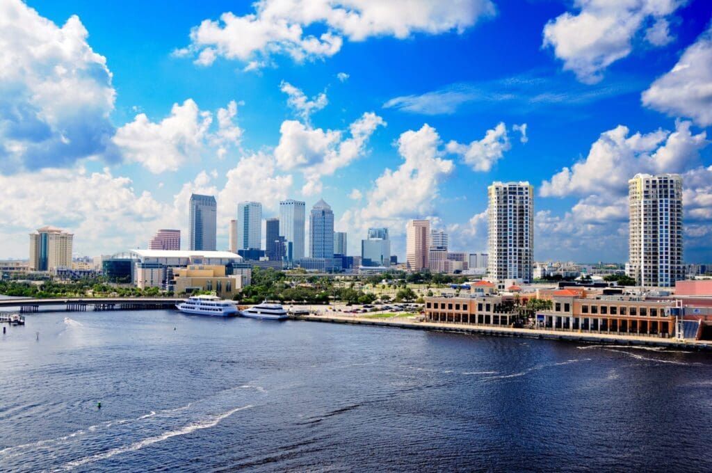 A view of a city skyline with tall modern buildings under a cloudy blue sky. A body of water is in the foreground, with yachts docked near the shore. The scene is vibrant and scenic.