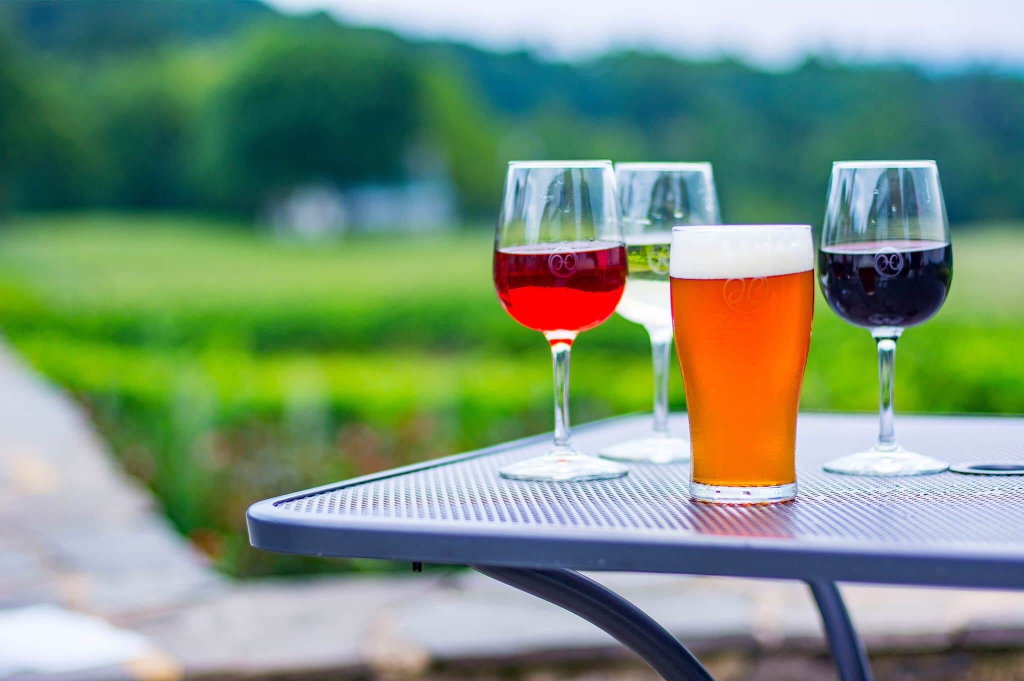 A metal table outdoors holds a pint of beer in the foreground, with three wine glasses in the background, containing red, white, and rosé wine. The lush, blurred landscape is green and inviting.