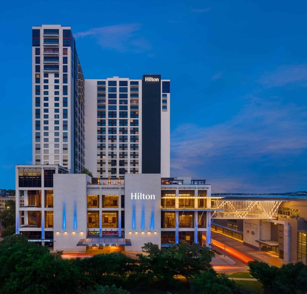 A tall hotel building with Hilton signage, featuring a modern design and numerous windows, stands against a twilight sky. Lights illuminate the lower facade with blue accents. Trees and a road with light trails are visible in the foreground.