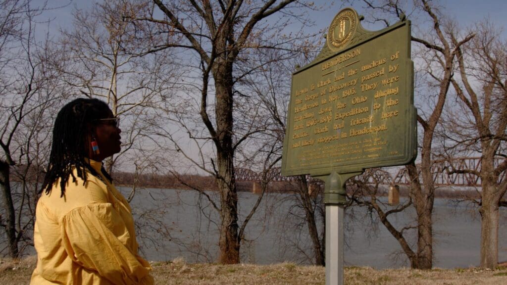 A person in a yellow jacket stands by a historical marker near a riverbank. The marker is surrounded by leafless trees, and a bridge is visible in the background.