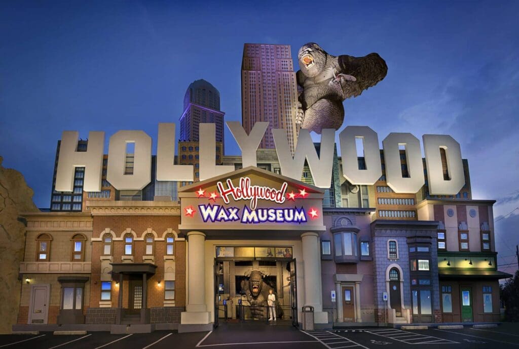Front view of Hollywood Wax Museum with large HOLLYWOOD letters and a giant gorilla figure perched atop. The building features various architectural styles and is illuminated against a twilight sky.