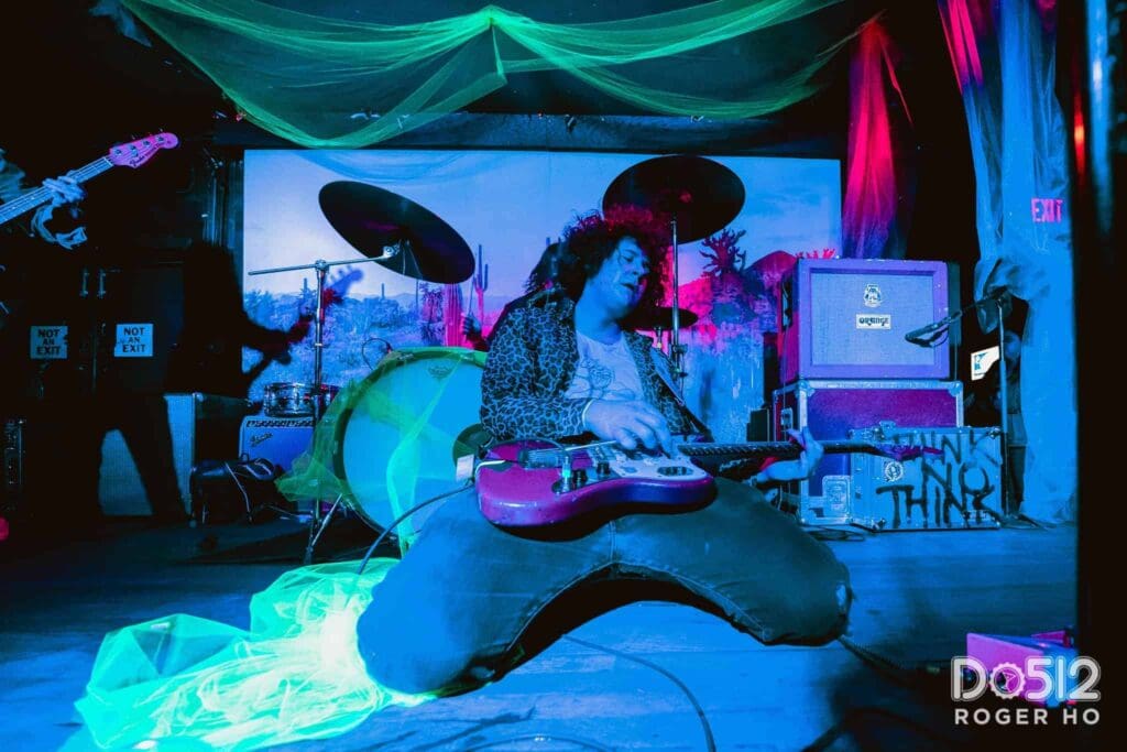 Musician kneels on stage playing an electric guitar energetically. Behind them, vibrant stage lights and a colorful backdrop create a lively atmosphere. Amplifiers and drums are visible, and a glowing green fabric accents the scene.
