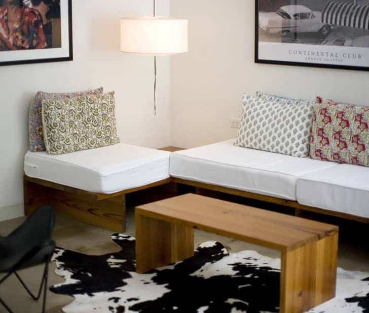 A cozy living room with a wooden L-shaped sofa featuring white cushions and patterned throw pillows. A wooden coffee table sits on a black and white cowhide rug. A floor lamp and framed wall art complete the decor.