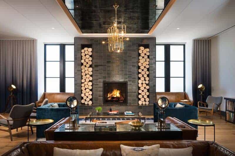 A modern living room features a central fireplace with logs stacked on each side. Two blue sofas and brown leather chairs encircle the fireplace. A large chandelier hangs from the ceiling, and tall windows illuminate the space.