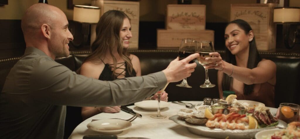 Three people sit at a restaurant table clinking wine glasses in a toast. The table is set with plates of oysters and shrimp. They are all smiling and appear to be enjoying their meal and company in a warmly lit setting.