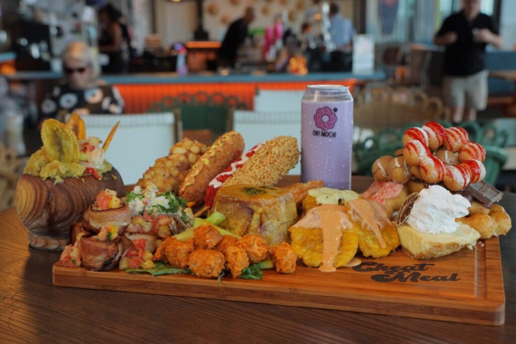 A wooden board is filled with various foods including fried items, sandwiches, pastries, and a can labeled ONE MOKO. A woman in the background sits at a table, wearing sunglasses and a patterned top.