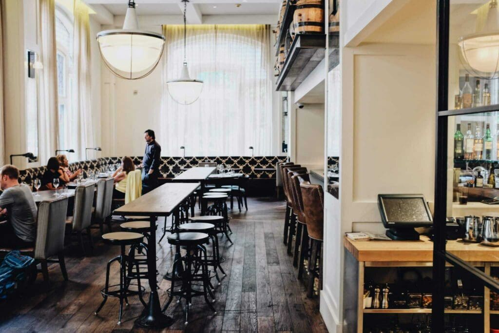 A modern, cozy restaurant interior with wooden floors. A long table with bar stools sits in the center. People are dining on the left, while the right side features a bar with stools and shelves of bottles. Large windows let in natural light.