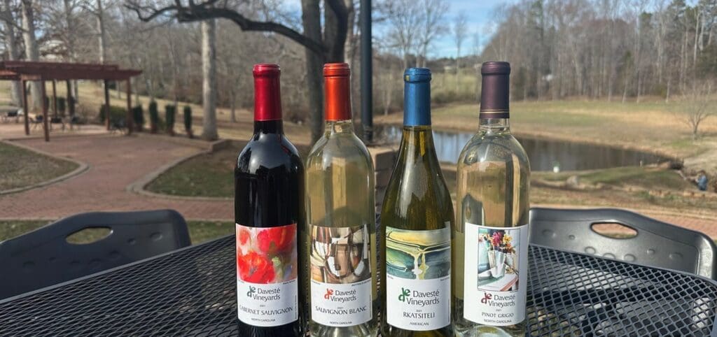 Four bottles of wine from different varieties (red and white) are displayed on a black metal table outdoors. The background features a pond, trees, and a brick pathway, creating a serene and picturesque setting.