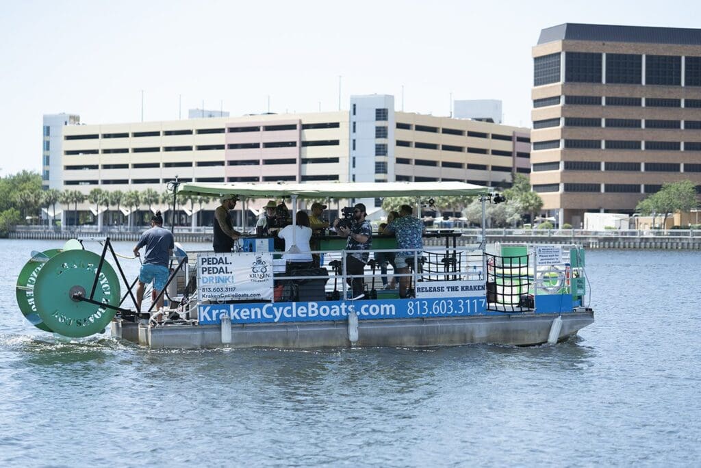A group of people enjoying a ride on a pedal-powered cycle boat named Kraken CycleBoats on a sunny day. The boat is on a calm body of water, and multi-story buildings are visible in the background.