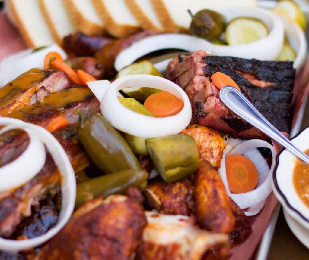 A close-up of a barbecue platter featuring ribs, grilled meat, onion rings, pickles, sliced carrots, and jalapeños. A spoon rests on the platter, and slices of toast are visible in the background.
