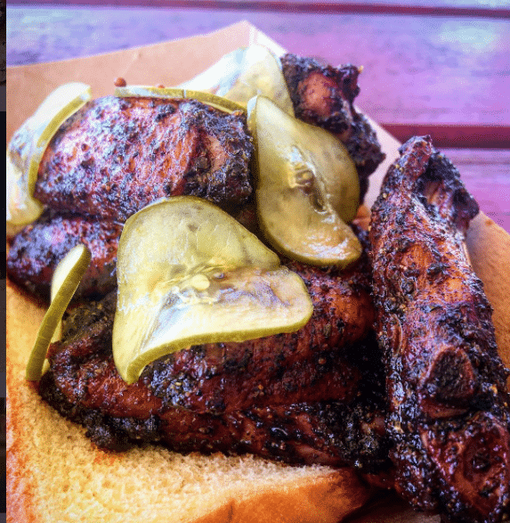 Grilled chicken wings topped with sliced pickles are served on a slice of bread, showcasing a rich, charred texture. The meal is presented on a wooden tray.