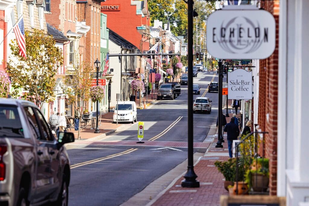 A quaint small-town street with brick buildings and shops on either side. Cars are parked along the street, and a few pedestrians are walking. Signs for local businesses, like Echelon and SideBar Coffee, are visible. American flags hang from buildings.