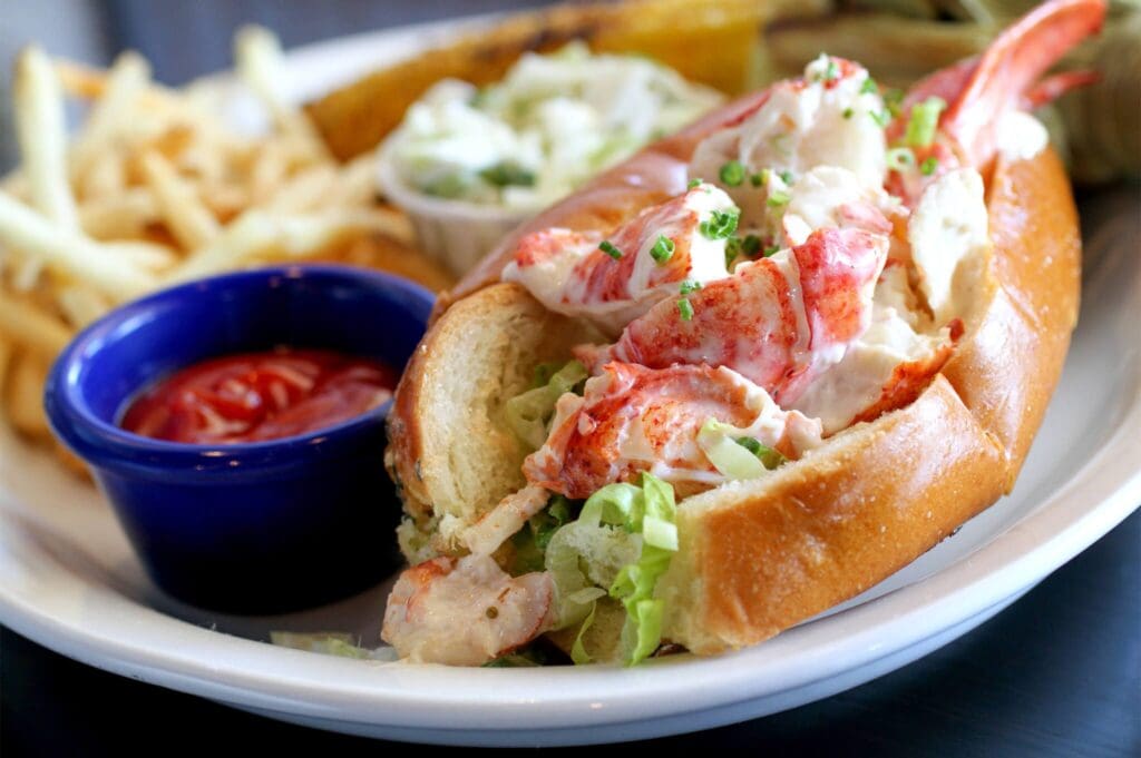 A lobster roll filled with lobster meat and garnished with chopped herbs, served on a plate with a side of crispy fries, coleslaw, and a small blue cup of ketchup. The roll is placed on a lightly toasted bun.