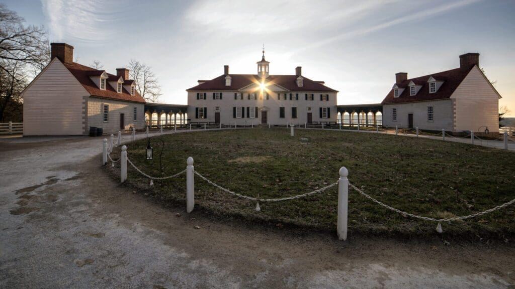 The image shows a historic white mansion with a red roof, featuring a semicircular layout and connected buildings. The sun is setting behind the central structure. A gravel path and a grassy area surrounded by a low rope fence are in the foreground.