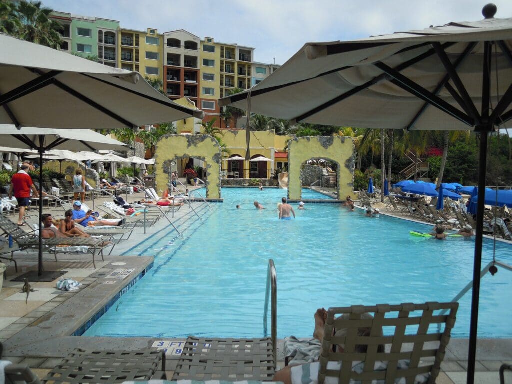 People relax by a hotel pool under large umbrellas. Some are swimming, while others lounge on sunbeds. The pool is surrounded by tropical plants and a multi-colored building in the background. The sky is partly cloudy.