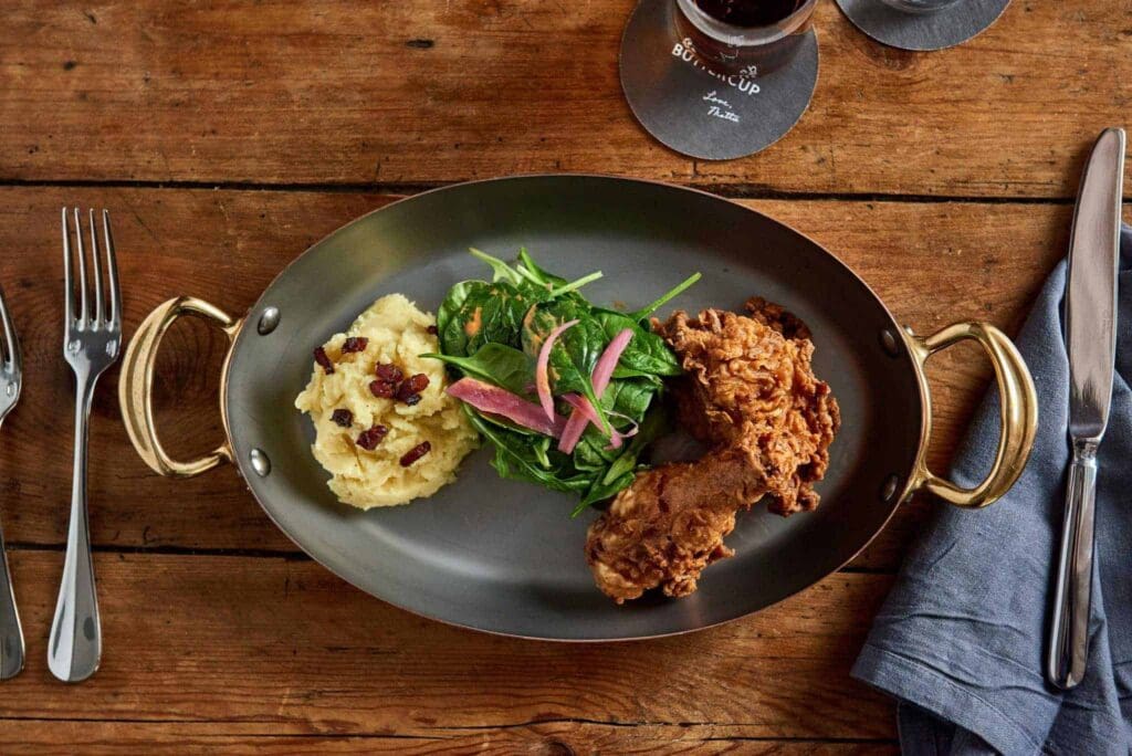 A plate featuring fried chicken, mashed potatoes with bacon bits, and a fresh salad with greens and pickled red onions. The plate sits on a wooden table with a fork, knife, and a blue napkin nearby.