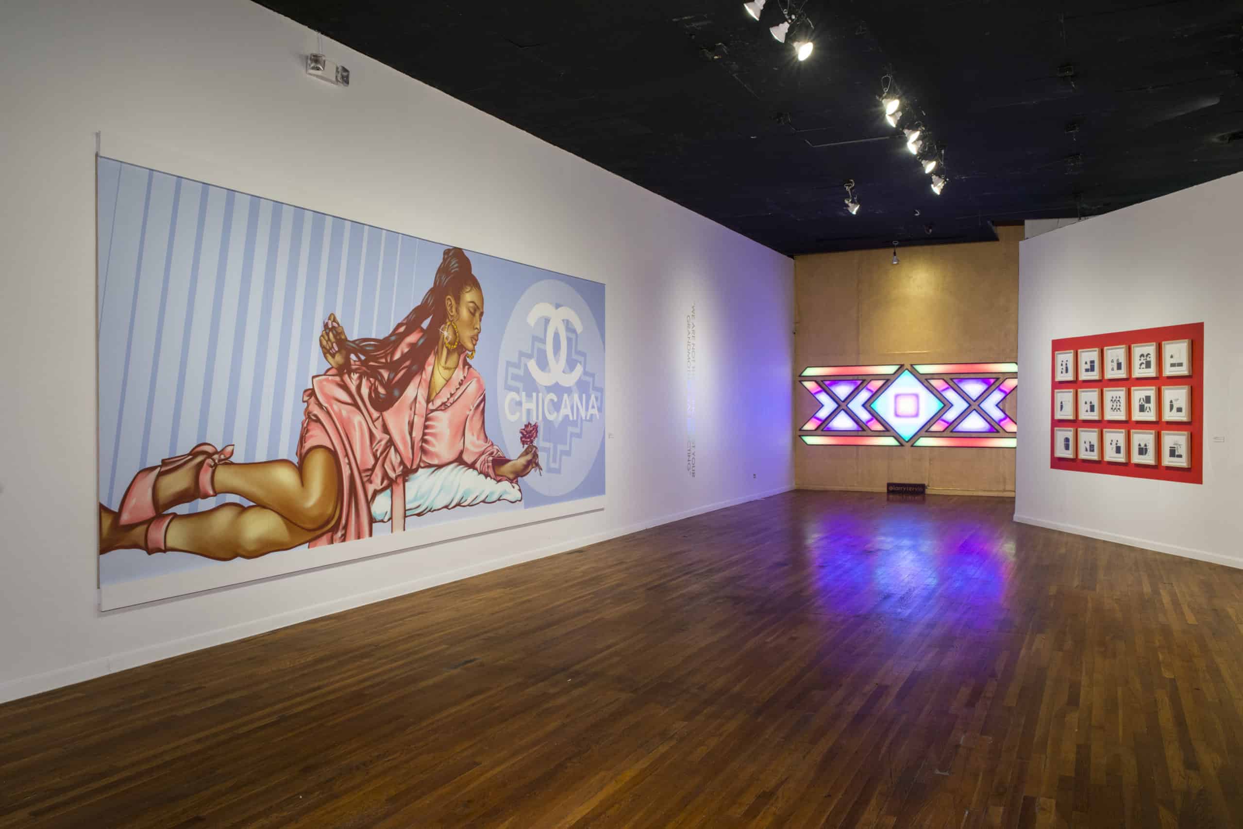 Art gallery with a large mural of a woman in pink, labeled Chicana, on the left wall. A colorful geometric light installation and a grid of small framed artworks are on the rear wall. The floor is wooden, and the ceiling is black.