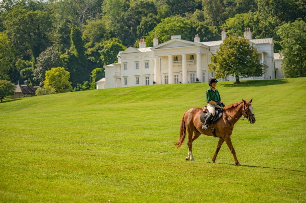 A person riding a brown horse on a vast green lawn in front of a large white mansion with pillars. The mansion is surrounded by trees under a clear blue sky.