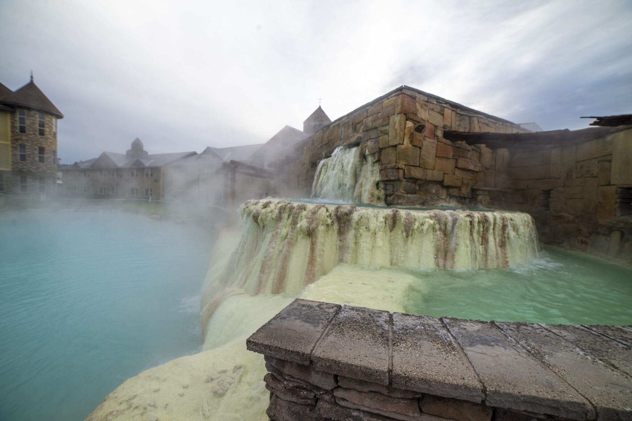 Mist surrounds a thermal hot spring with stone structures and green mineral deposits. Water cascades over the edge. In the background, cloudy skies and buildings with tall windows and pointed roofs are visible.