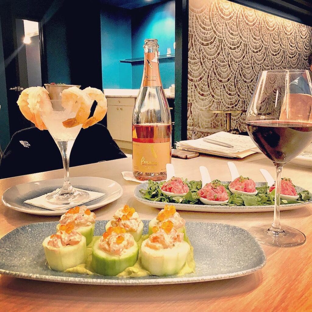 A table set with gourmet dishes: cucumber-wrapped appetizers topped with orange garnish, spoonfuls of pink tartare on greens, a glass of fried rings, a glass of red wine, and a bottle of rosé champagne against a decorative backdrop.