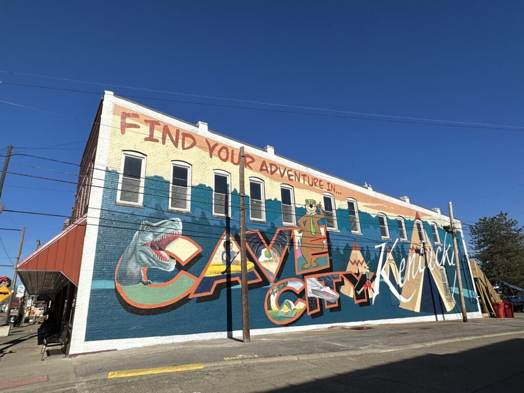 A colorful mural on the side of a building reads Find your adventure in Cave City, Kentucky. The artwork includes a dinosaur and vibrant lettering, set against a clear blue sky.