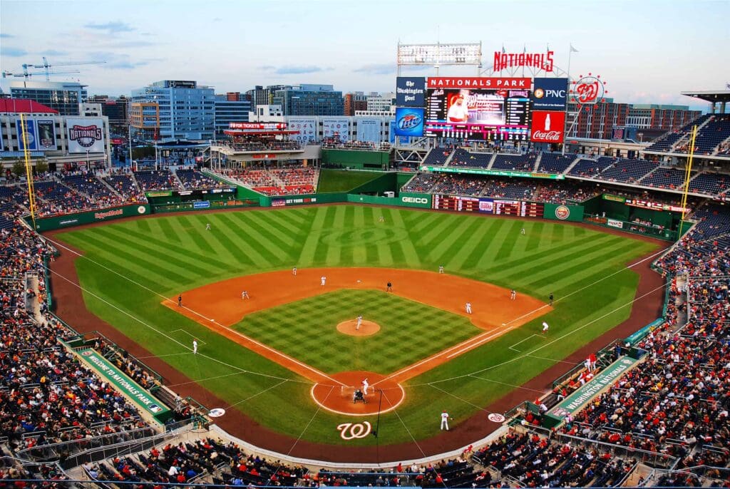 Aerial view of Nationals Park during a baseball game. The diamond is surrounded by lush green grass, with players on the field and a batter at home plate. The stands are partially filled with spectators, and the city skyline is visible in the background.