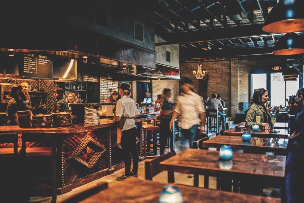 A lively restaurant interior with wooden decor and dim lighting. Staff are working behind a counter, while patrons stand and converse. Tables are adorned with small blue candles, and a chandelier hangs from the ceiling.