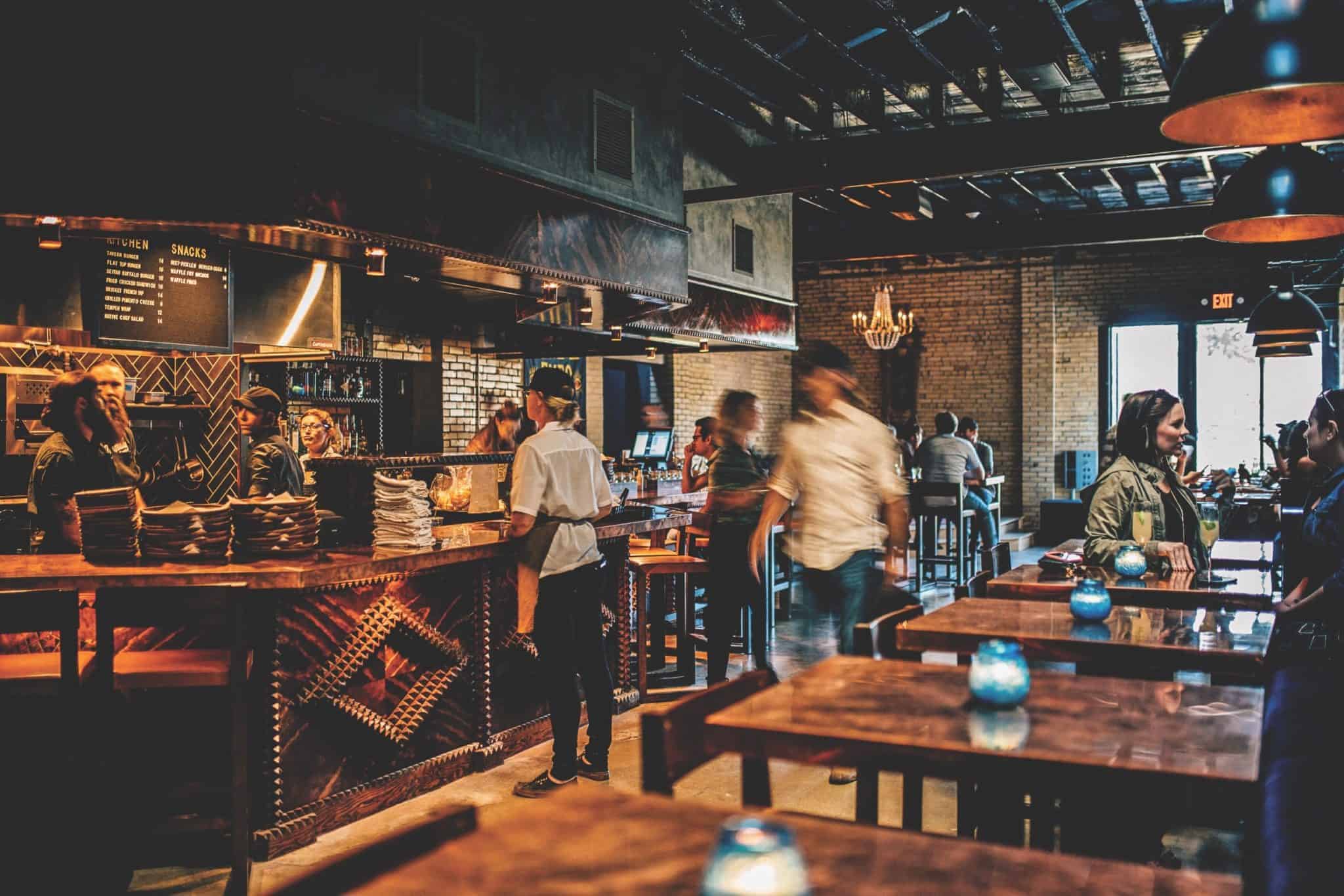 A lively restaurant interior with wooden decor and dim lighting. Staff are working behind a counter, while patrons stand and converse. Tables are adorned with small blue candles, and a chandelier hangs from the ceiling.