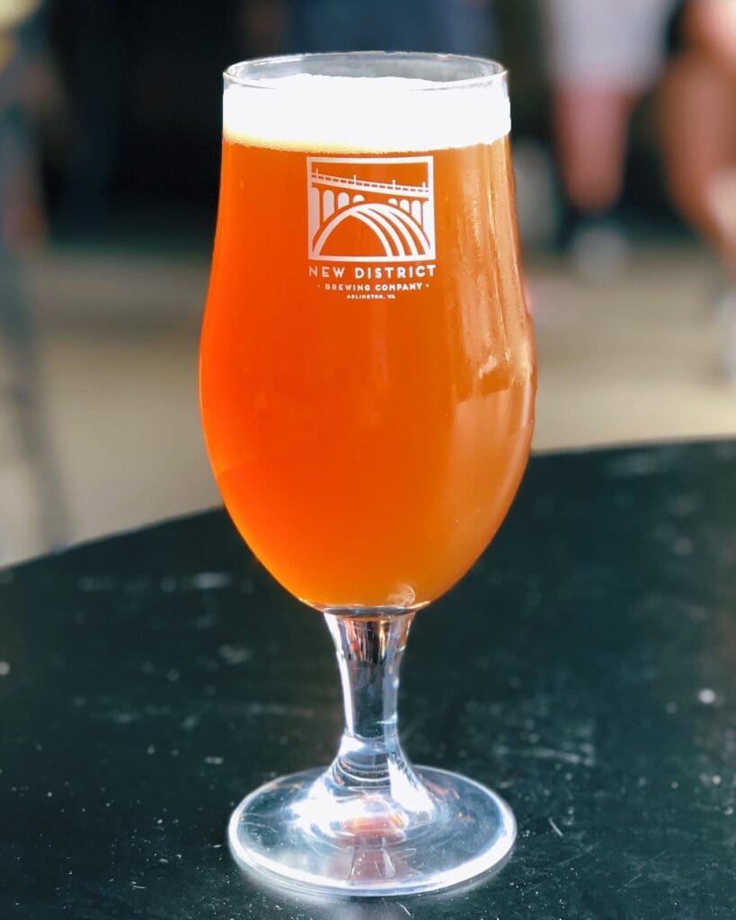 A frothy, amber-colored beer in a tulip glass with the logo of New District Brewing Company on it, placed on a black table in a dimly lit setting.
