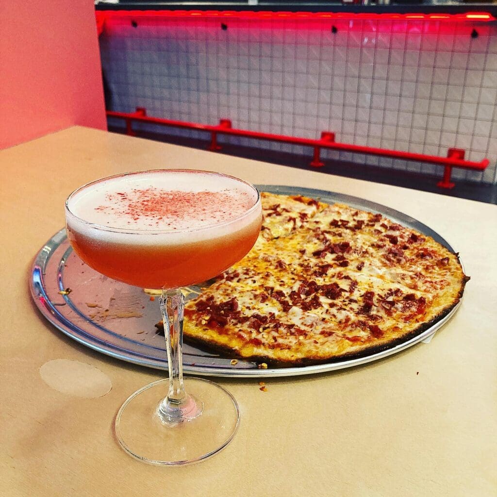 A cocktail with a pink frothy top in a coupe glass sits on a wooden counter. Next to it, a thin-crust pizza topped with cheese and bacon is on a metal tray. A white tiled wall with red accents is in the background.