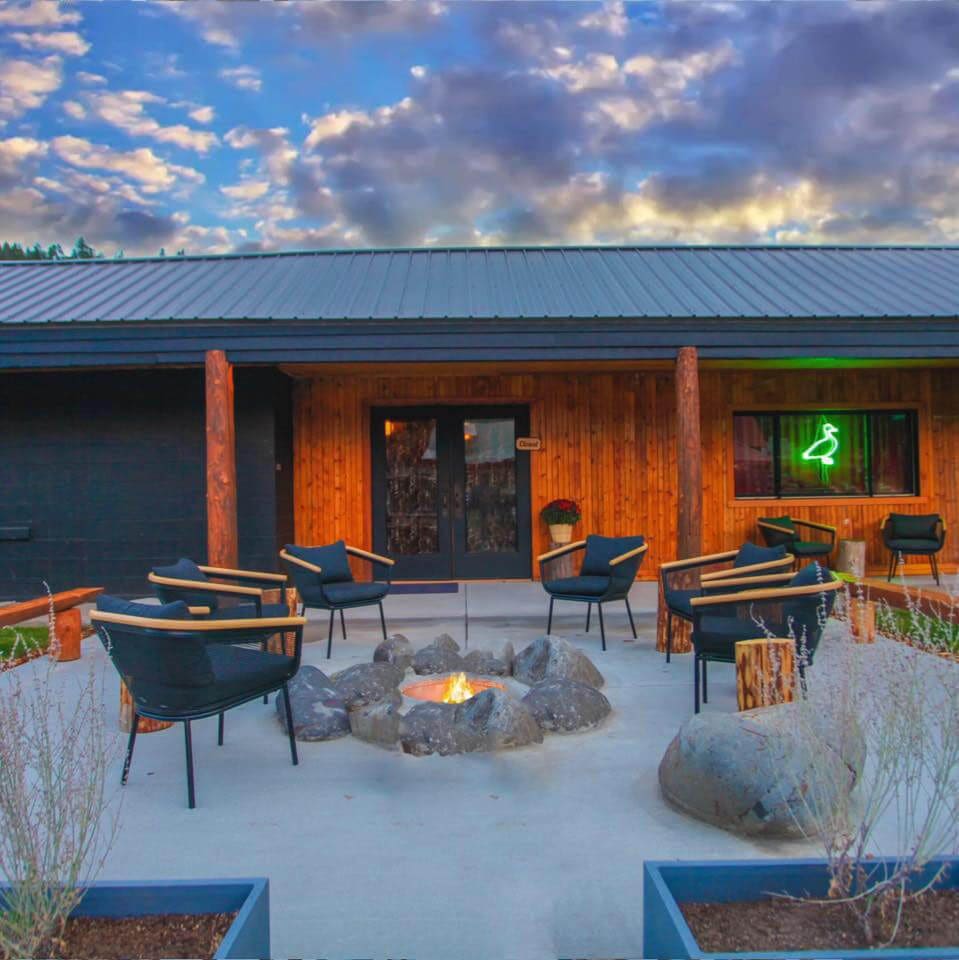 Outdoor seating area with black chairs surrounds a rock fire pit on a concrete patio. A building with wooden accents features a green neon light in the window. The sky is filled with clouds, reflecting colors of the setting sun.