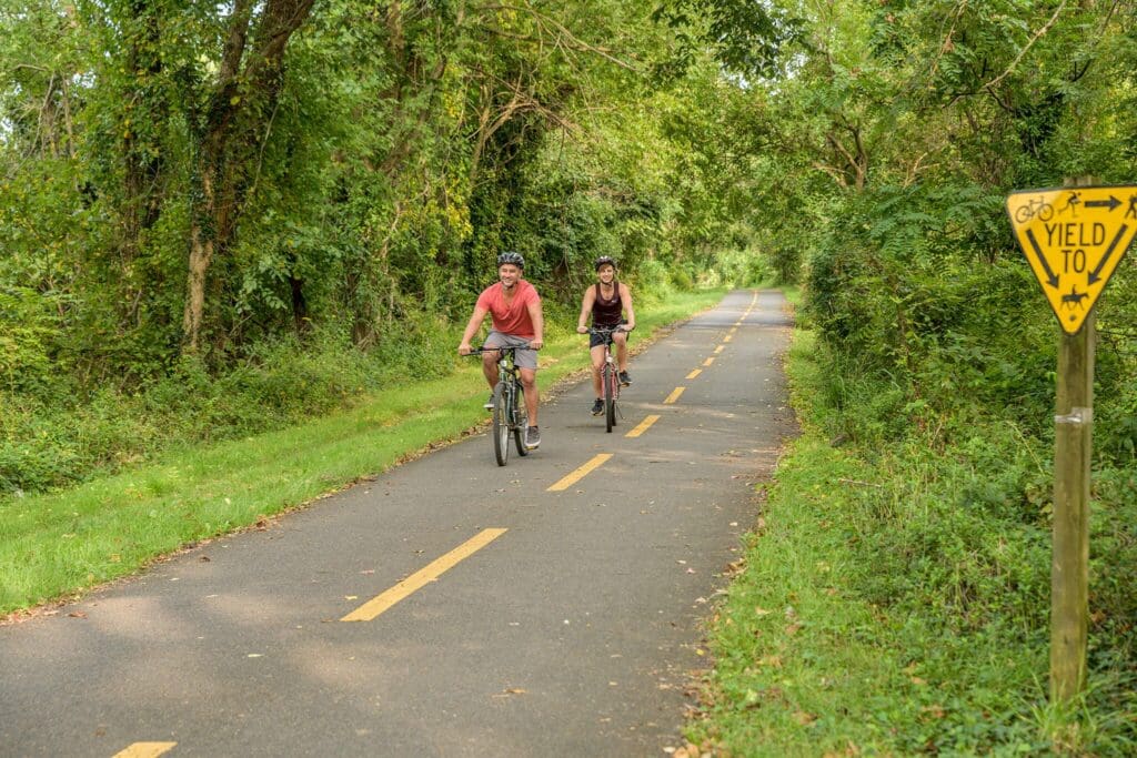 Two people ride bicycles along a tree-lined path with a yellow dashed line. A yellow sign indicates Yield to with icons of pedestrians and bikers. The scene is green and lush, suggesting a park-like setting.