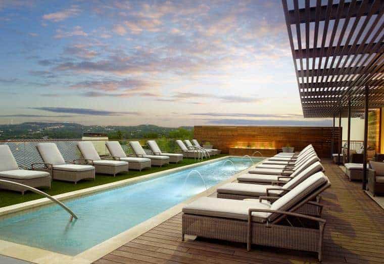 A modern rooftop pool with multiple lounge chairs under a clear sky. The pool features water jets, surrounded by a deck with wooden pergola accents. Scenic views of hills are visible in the distance as the sun sets.