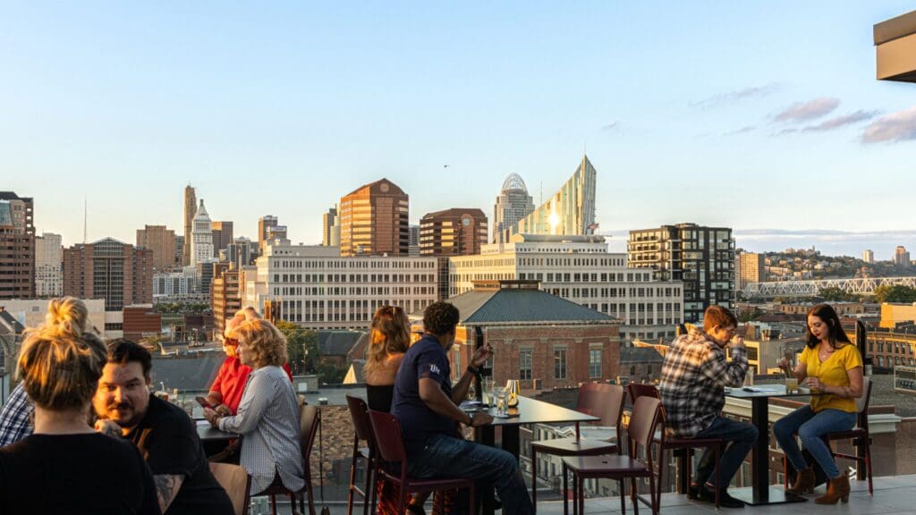 People are dining on a rooftop patio with tables and chairs. In the background, a city skyline is visible under a clear blue sky. The scene suggests a casual, social atmosphere in an urban setting.