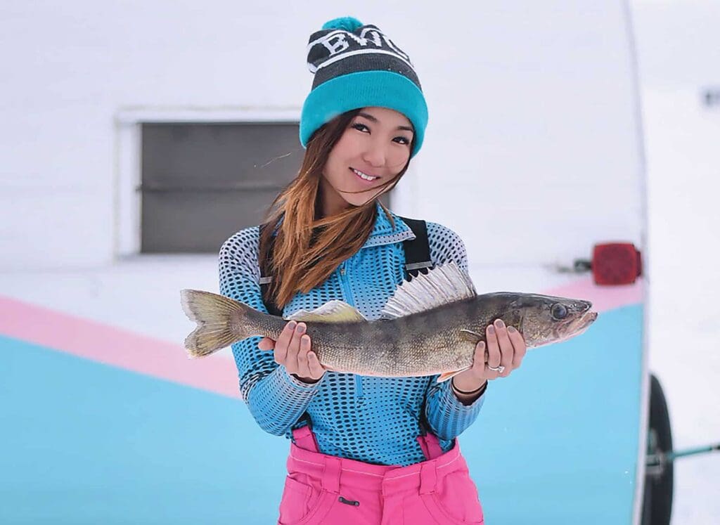 Woman wearing a blue beanie and pink overalls holds a fish with a small smile, standing in front of a light-colored, partially visible trailer or vehicle. The scene suggests a fishing outing in a snowy environment.