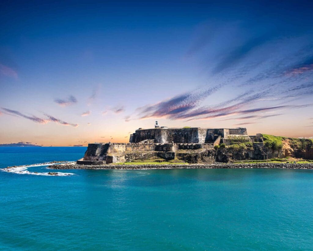 A historic fortress sits on a rocky coastline, surrounded by bright blue ocean under a clear sky with scattered clouds. The structure is made of weathered stone, providing a dramatic contrast to the vibrant seascape.