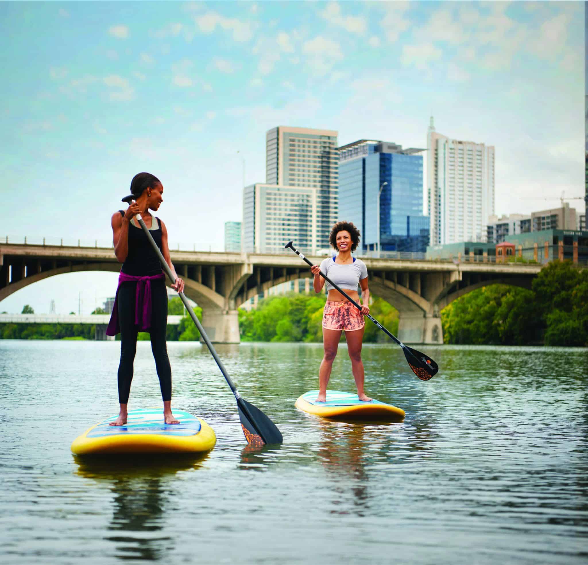 Two people are paddleboarding on a calm river with a cityscape in the background. A bridge stretches across the river, and the skyline features tall modern buildings. The sky is partly cloudy, creating a serene atmosphere.
